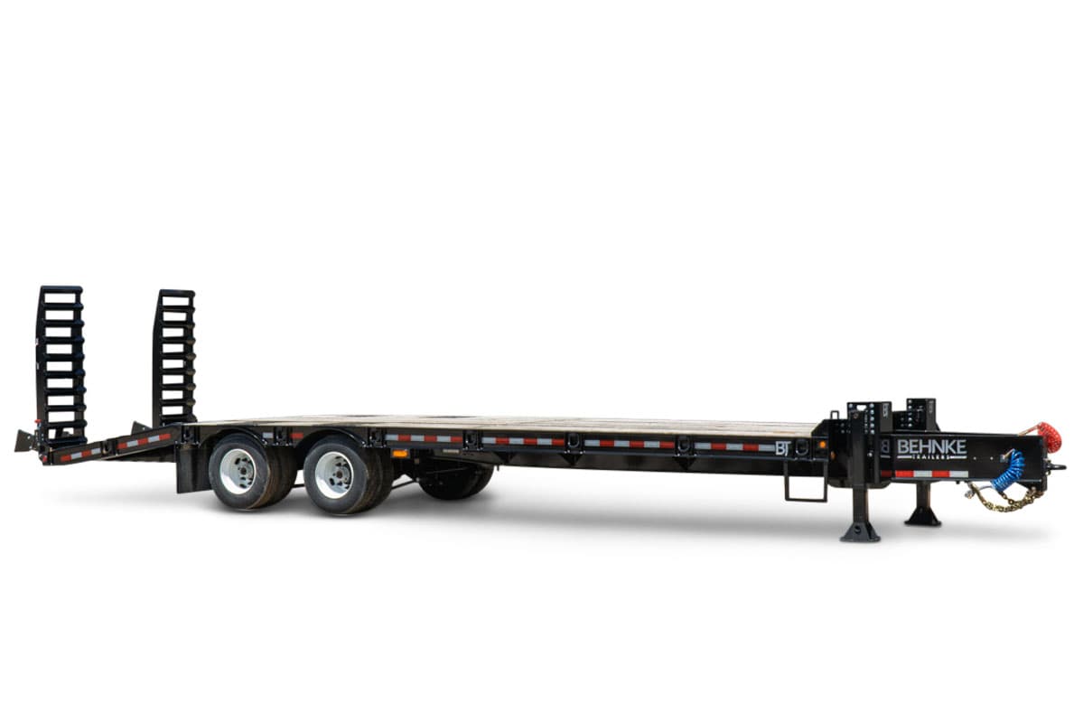2026 Behnke FB8X30SDPT-40L flatbed equipment trailer, black steel deck, dual tandem axles, fold-up ramps, pintle hitch, side-angle view