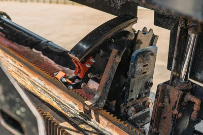 Ditch Witch JT20 carriage assembly close-up showing rack-and-pinion drive, vise clamp, hydraulic cylinder, and drill pipe with wear from use.