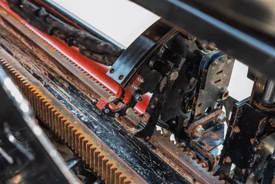 Ditch Witch JT20 carriage rack-and-pinion thrust system closeup, showing toothed drive rack, vise jaw, and red drill pipe with field wear.