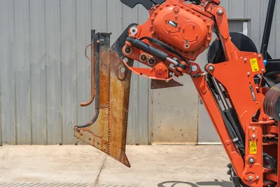 2024 Ditch Witch RT125Q VP120G vibratory plow attachment close-up, worn steel blade, orange hydraulic arm, safety decals visible