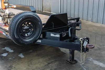 2023 MAXX-D C6X mud trailer tongue detail showing toolbox, pintle hitch, safety chains, jack stand, and radial ST tire on black steel wheel