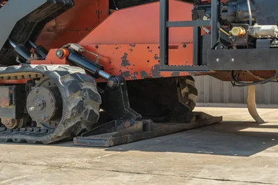 2014 Ditch Witch JT100 Mach 1 undercarriage close-up showing rubber track, drive sprocket, hydraulic cylinder, and anchor plate with worn paint
