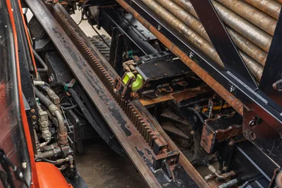 2016 Ditch Witch JT25 drill frame closeup showing rack-and-pinion carriage, vise assembly, hydraulic lines, and loaded drill pipe rack