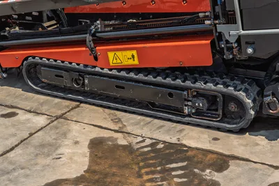 2023 Ditch Witch JT20 rubber track undercarriage close-up, electrical hazard warning decal visible on orange frame, hydraulic lines above