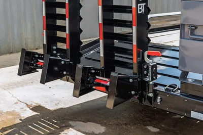2026 Behnke CBCT2324E-BT-R trailer rear closeup showing dual pipe rack uprights, stabilizer jacks, red reflective tape, and latch hardware