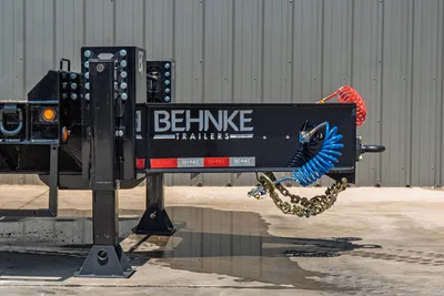 Behnke FB8X30SDPT-40L trailer tongue closeup showing gooseneck hitch, coiled air lines, safety chains, jack stands, and reflective markings.