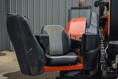 Ditch Witch JT24 operator seat close-up, gray vinyl cushion with logo, worn armrests, orange frame, engine compartment visible behind
