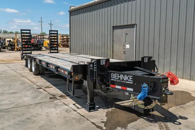 2026 Behnke FB8X30SDPT-40L flatbed trailer, rear 3/4 view, black steel deck, dual axles, coiled air lines, safety chains visible